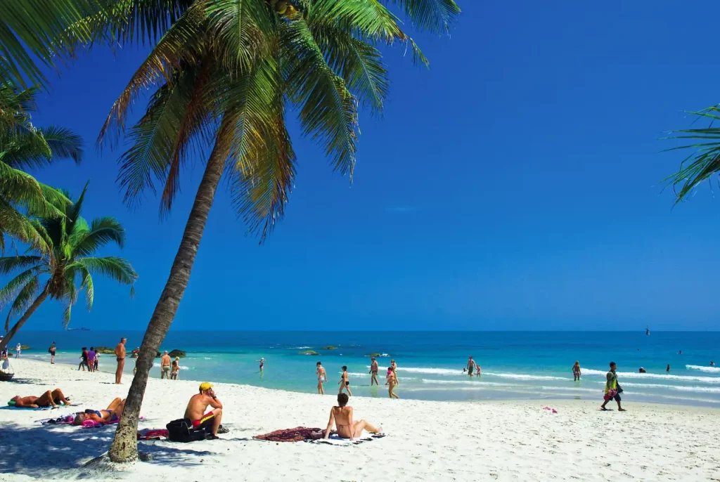 Blauer Himmel und strahlend weißer Strand von Pranburi, Thailand, mit Palme und Urlaubern.